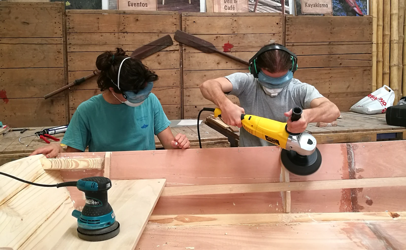 Dos chicos con protectores auditivos y barbijo lijan una tabla junto al casco de un bote en el taller